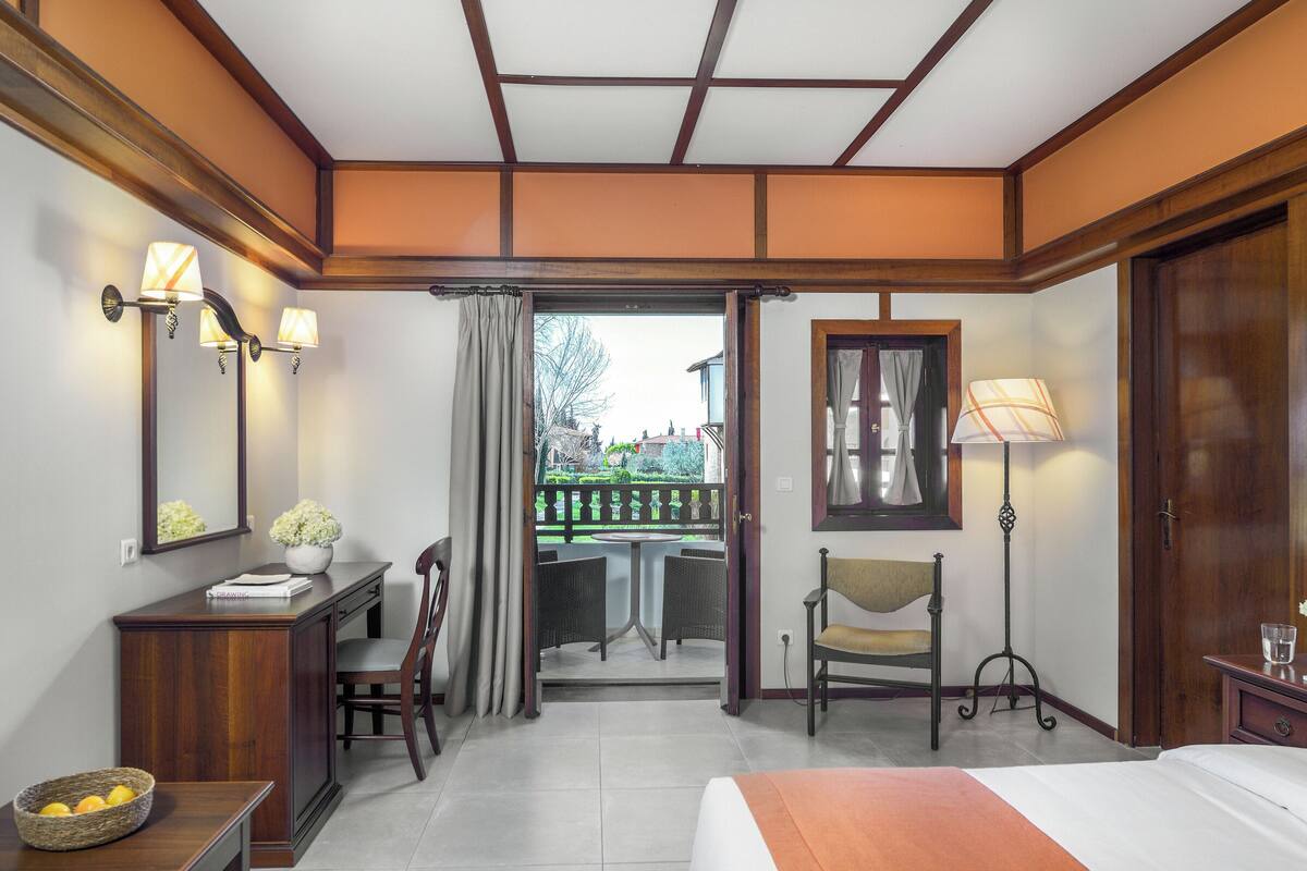 Ein Hotelzimmer mit einem Bett, einem Schreibtisch mit Lampe, einem Sessel, einem kleinen Tisch mit Vase und Blick auf den Balkon.