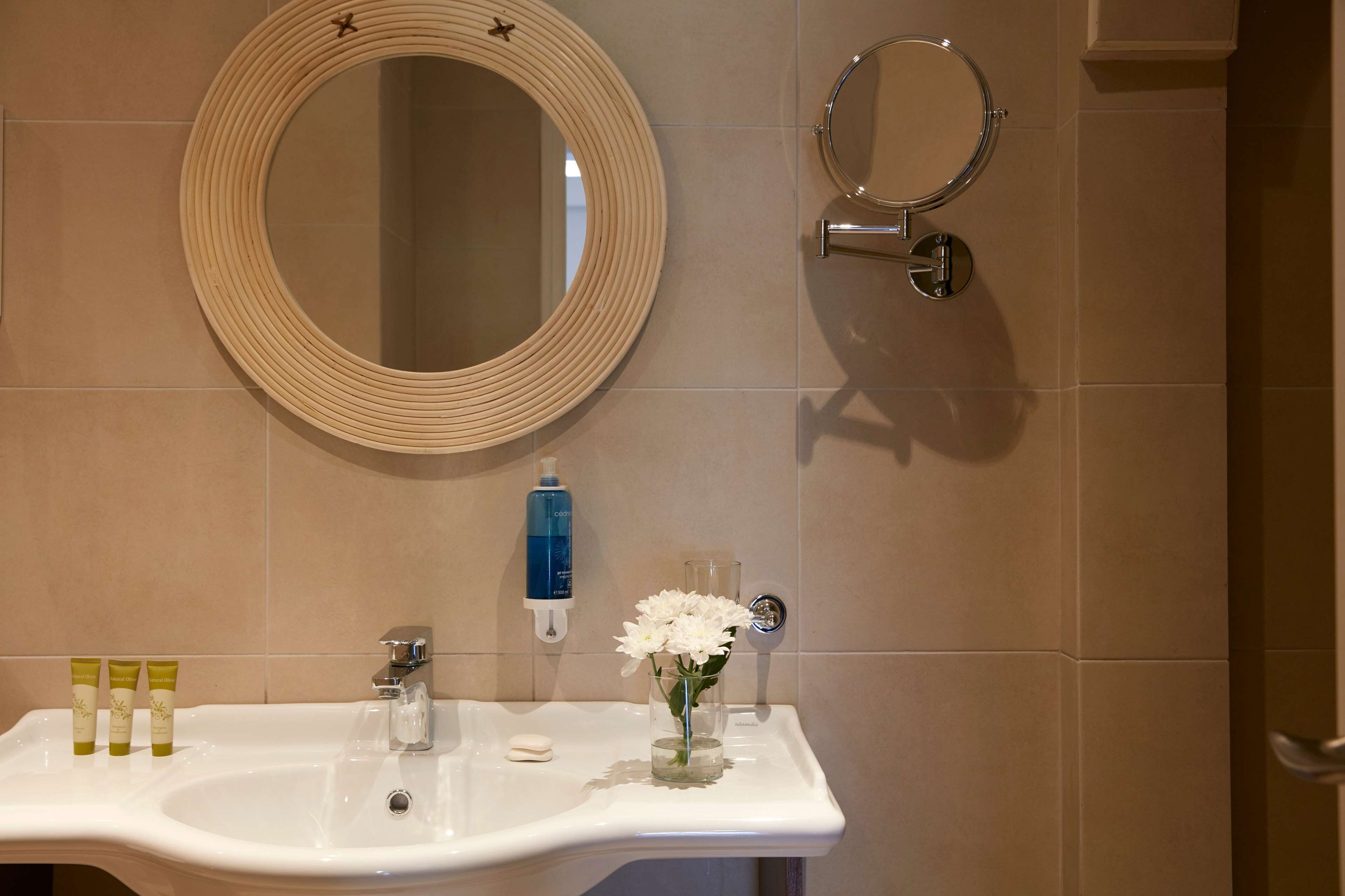 A bathroom with a round mirror, a sink with toiletries, and a glass of flowers.