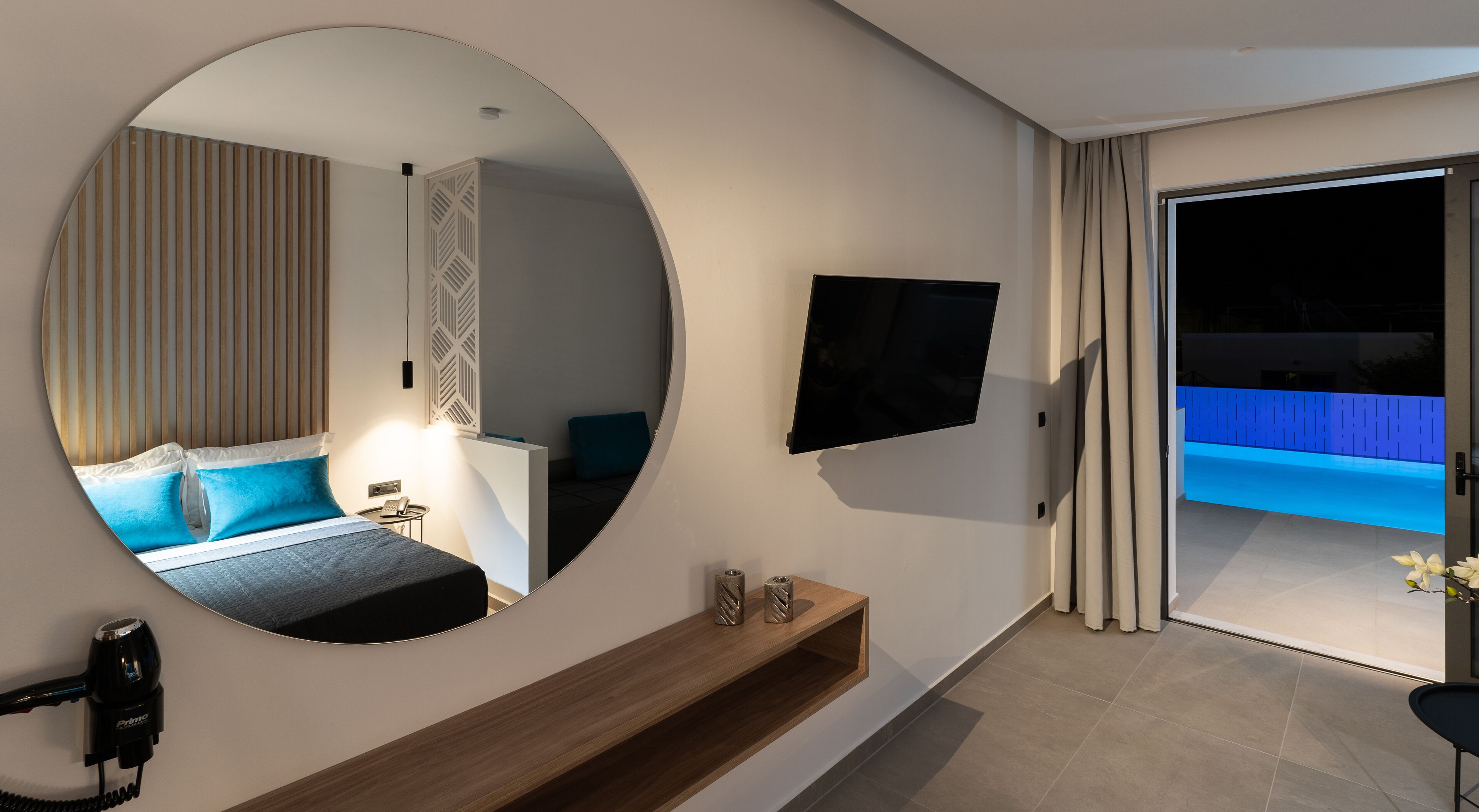 Ein modernes Hotelzimmer mit einem großen runden Spiegel, einem Flachbildfernseher, einem Holztischchen und Blick auf einen Poolbereich.