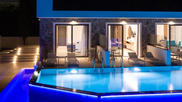 A modern poolside area with a swimming pool, lounge chairs, and a stone facade building with glass doors leading to a room. - Zoes Hotel (Rhodes)