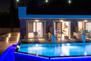 A modern poolside area with a swimming pool, lounge chairs, and a stone facade building with glass doors leading to a room. - Zoes Hotel (Rhodes)