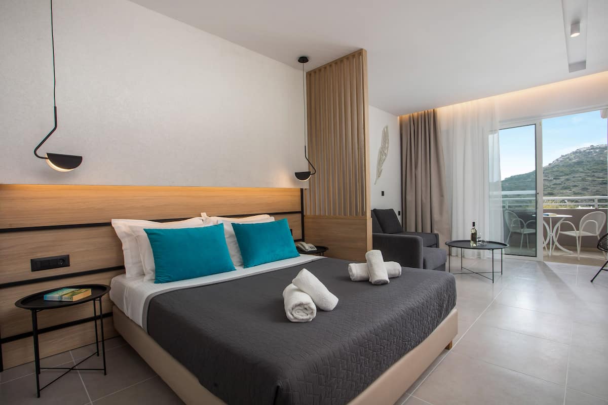 A modern hotel room with a large bed, a small side table, a desk, and a balcony with a view.