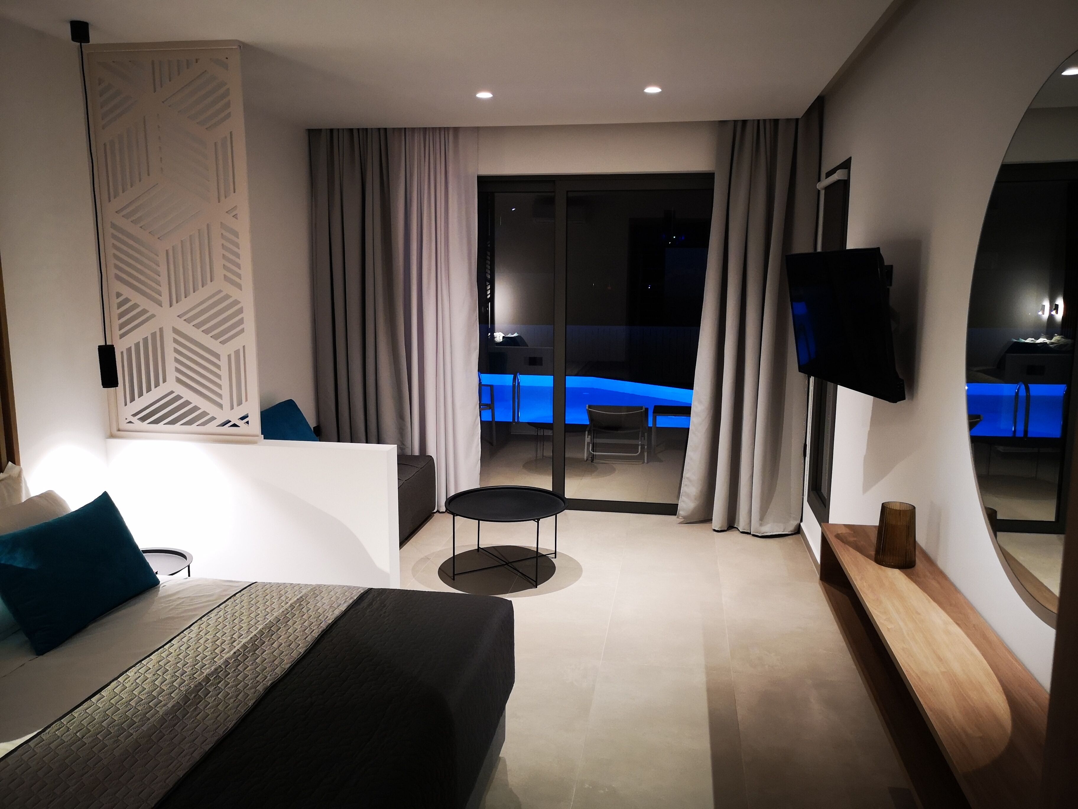 A modern hotel room with a bed, a small table, a TV, and a balcony with a pool.