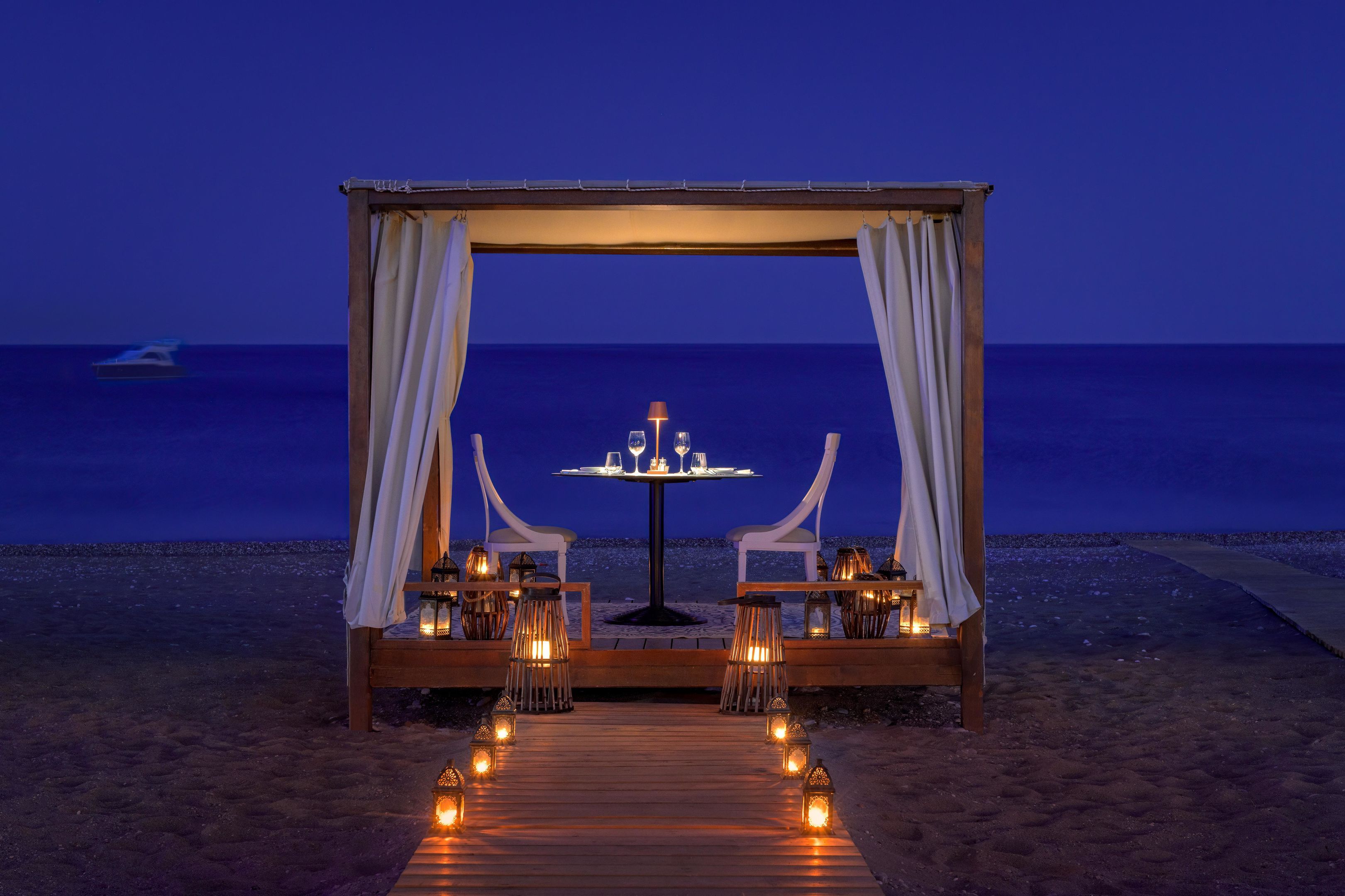 Decoración romántica en la playa con una mesa y sillas de madera, velas y vista al océano de noche.