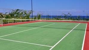 A tennis court with a net, surrounded by a fence, and a view of the ocean. - Rodos Palladium Leisure & Wellness (Rhodes)
