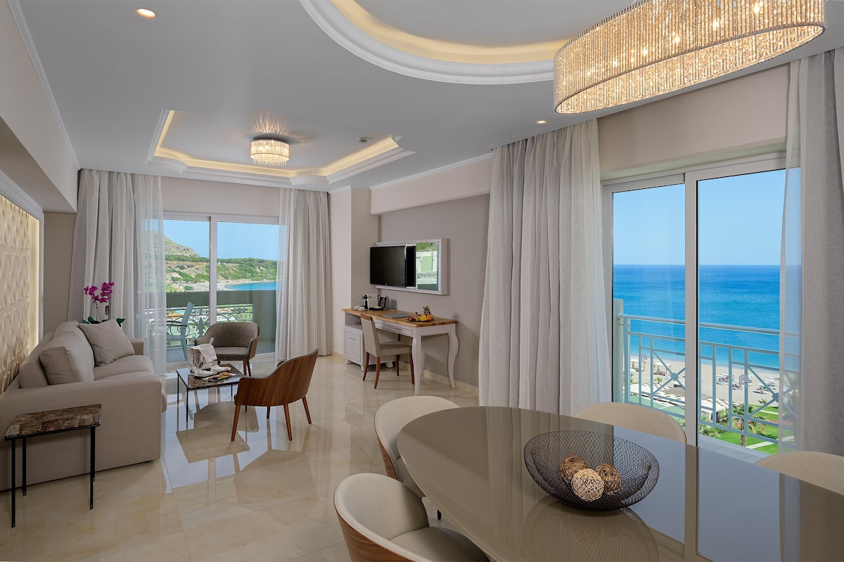 A modern living room with a large dining table, a sofa, and a TV. The room has large windows with a view of the ocean.