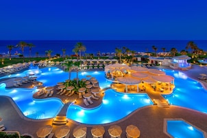 Uma área de piscina em resort com várias piscinas, espreguiçadeiras, guarda-sóis e vista para o oceano à noite.