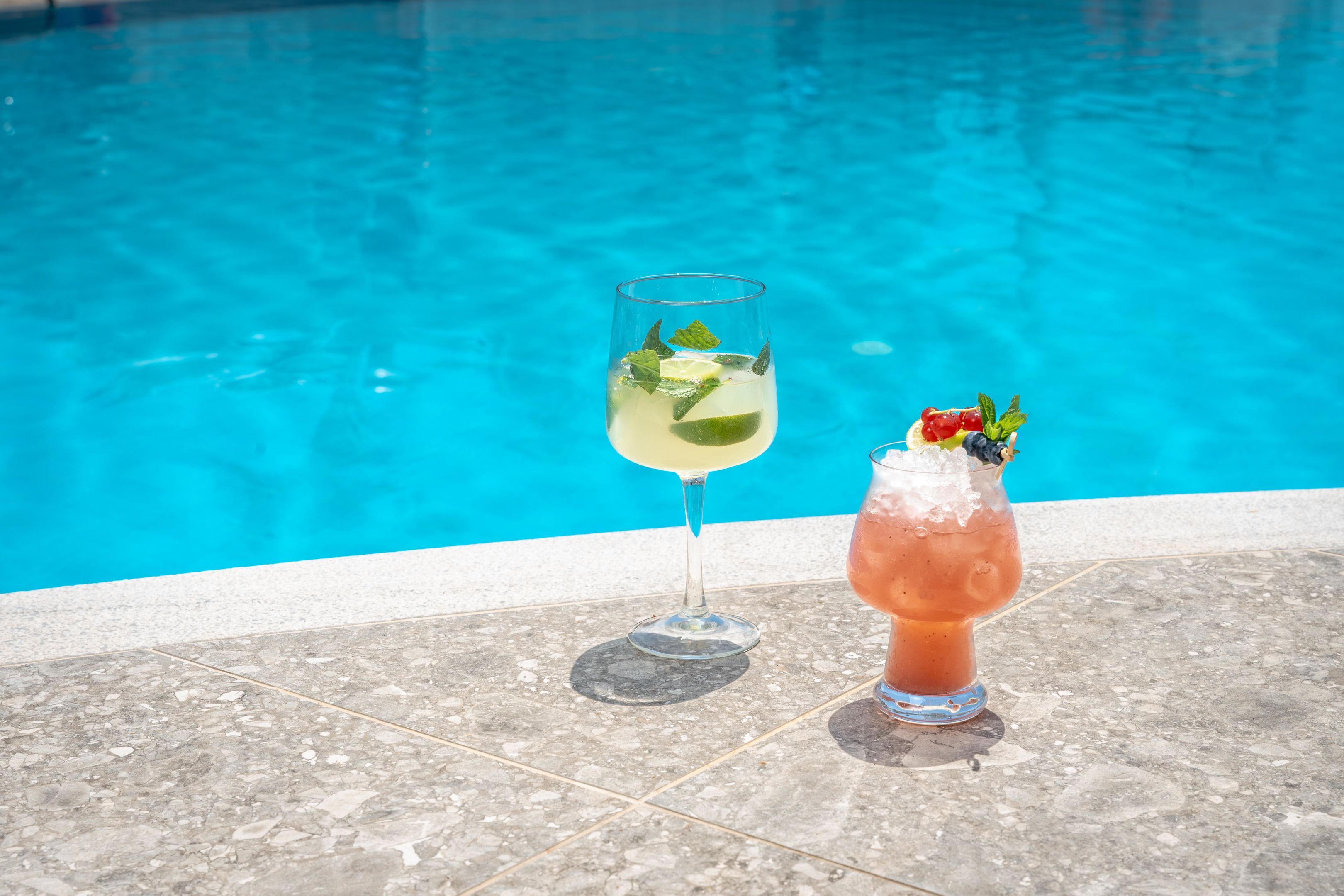 Dos bebidas sobre una superficie de baldosas junto a una piscina.