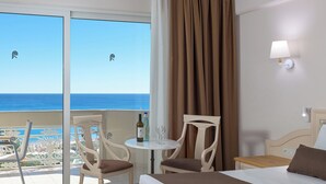 A hotel room with a bed, a table with wine bottles, two chairs, a view of the ocean, and a balcony with a railing. - Rodos Palladium Leisure & Wellness (Rhodes)