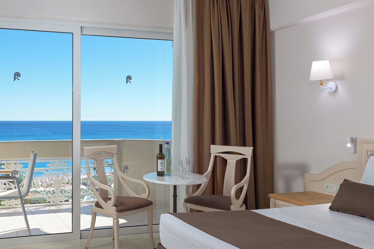 A hotel room with a bed, a table with wine bottles, two chairs, a view of the ocean, and a balcony with a railing.