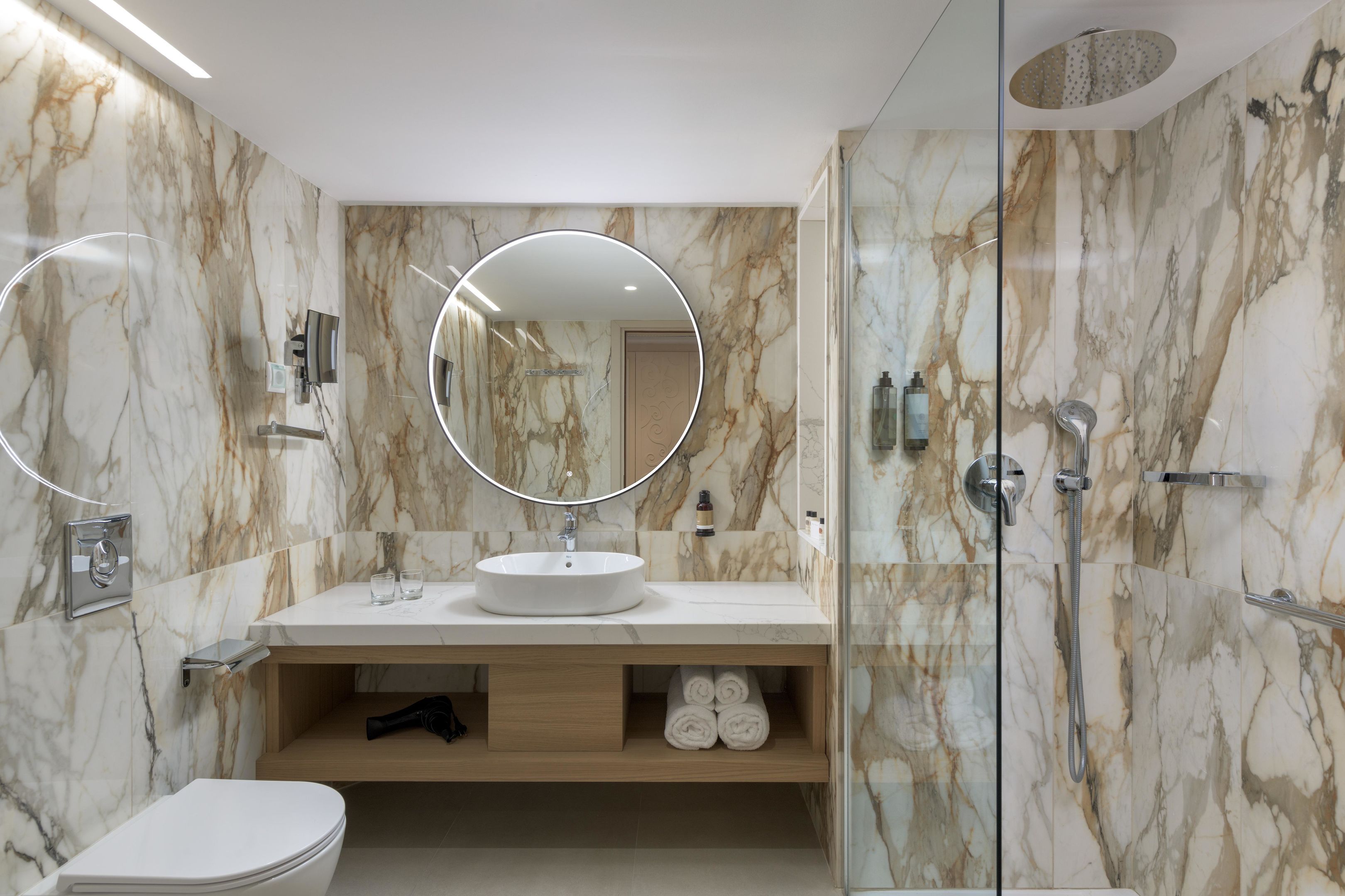A modern bathroom with a marble vanity, a round mirror, a toilet, and a glass-enclosed shower area.