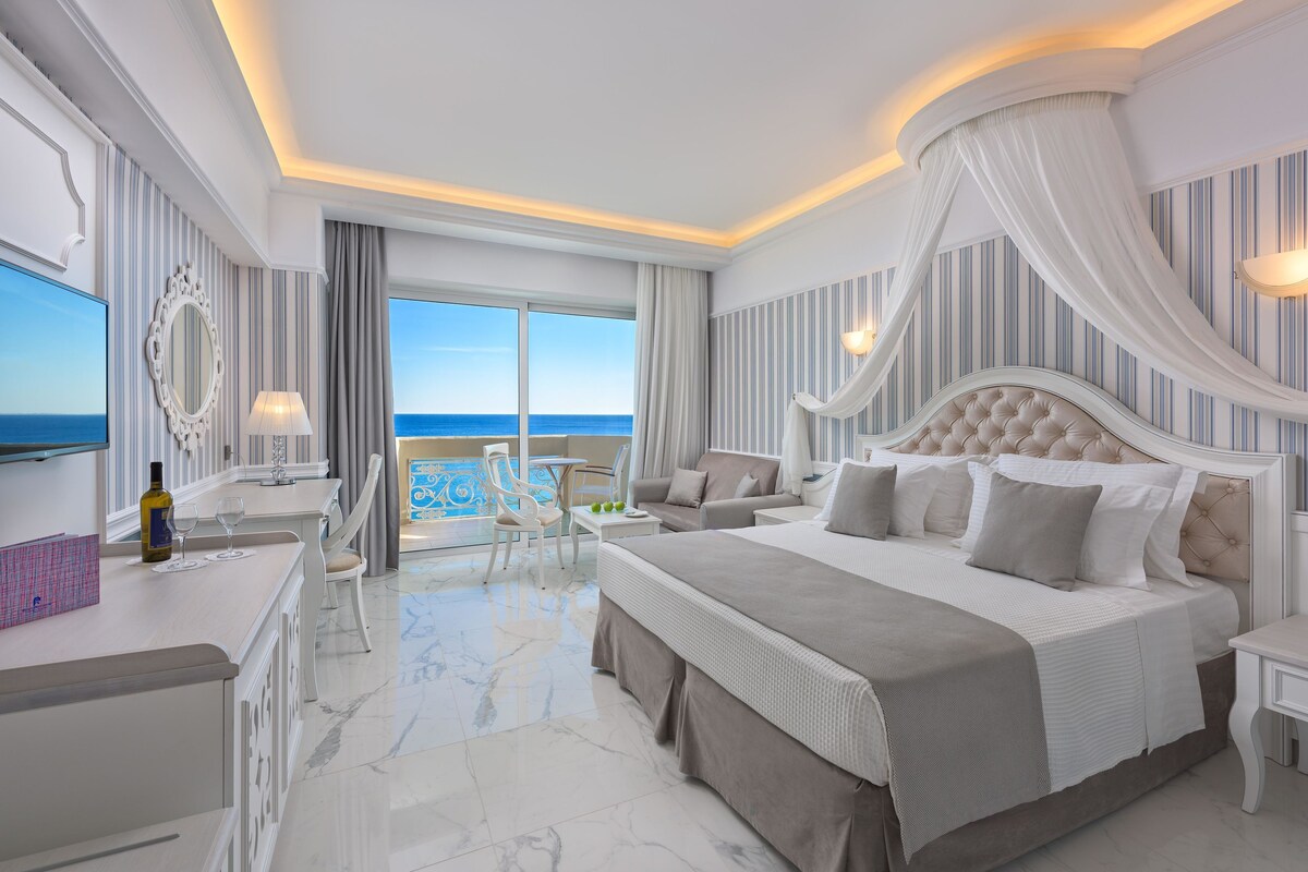 A spacious bedroom with a large bed, a sitting area with a view of the sea, and a balcony.
