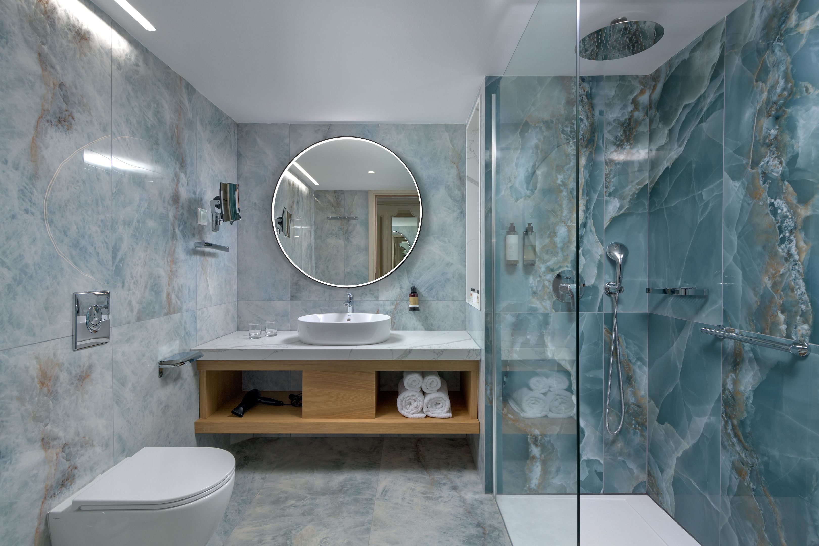Un baño moderno con lavamanos de mármol, un espejo redondo, cabina de ducha con mampara de vidrio y un inodoro.