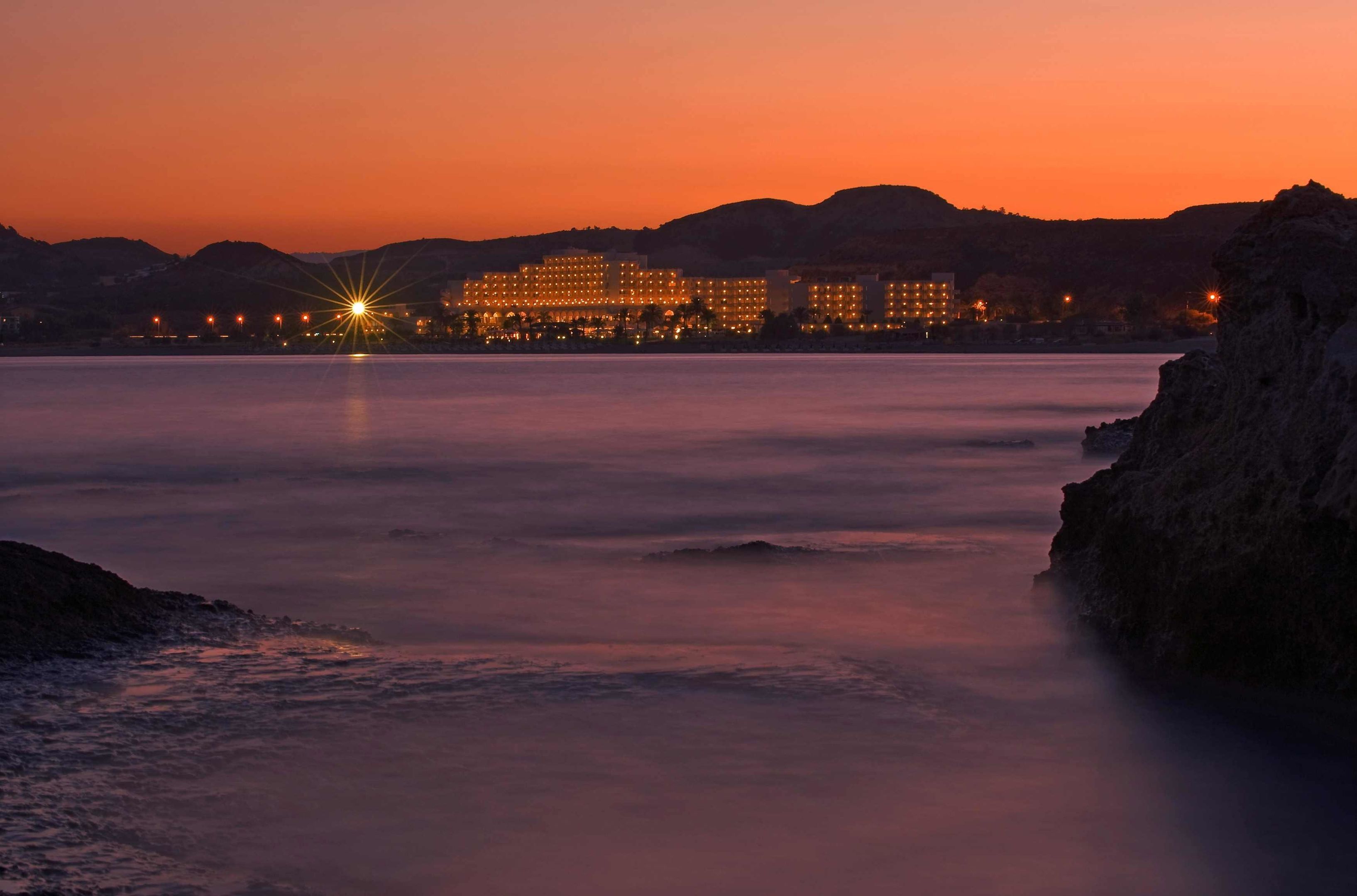 Un hotel costero con un edificio grande, agua tranquila y un atardecer.