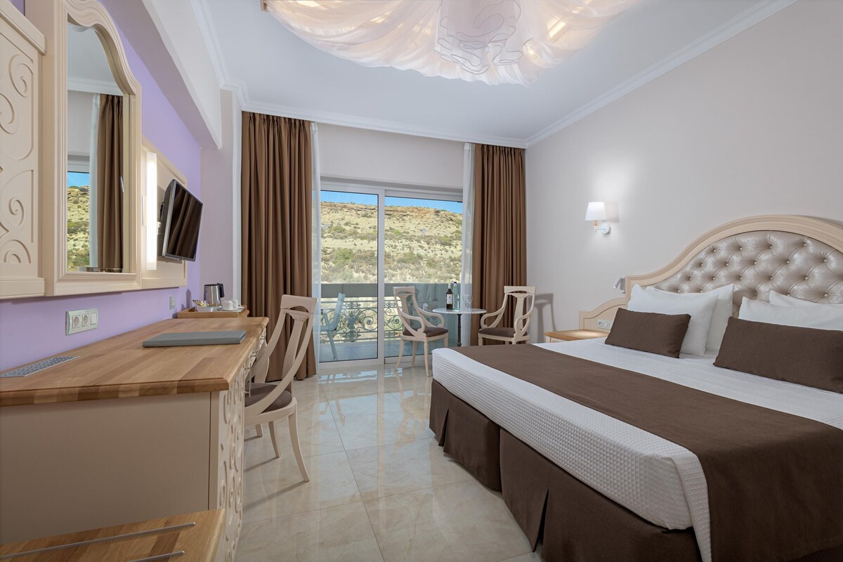 A hotel room with a bed, a desk, a chair, a TV, and a balcony with a view of the hills.