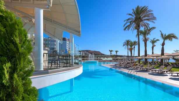 A pool area with lounge chairs, palm trees, and a building with a curved roof. - Rodos Palladium Leisure & Wellness (Rhodes)