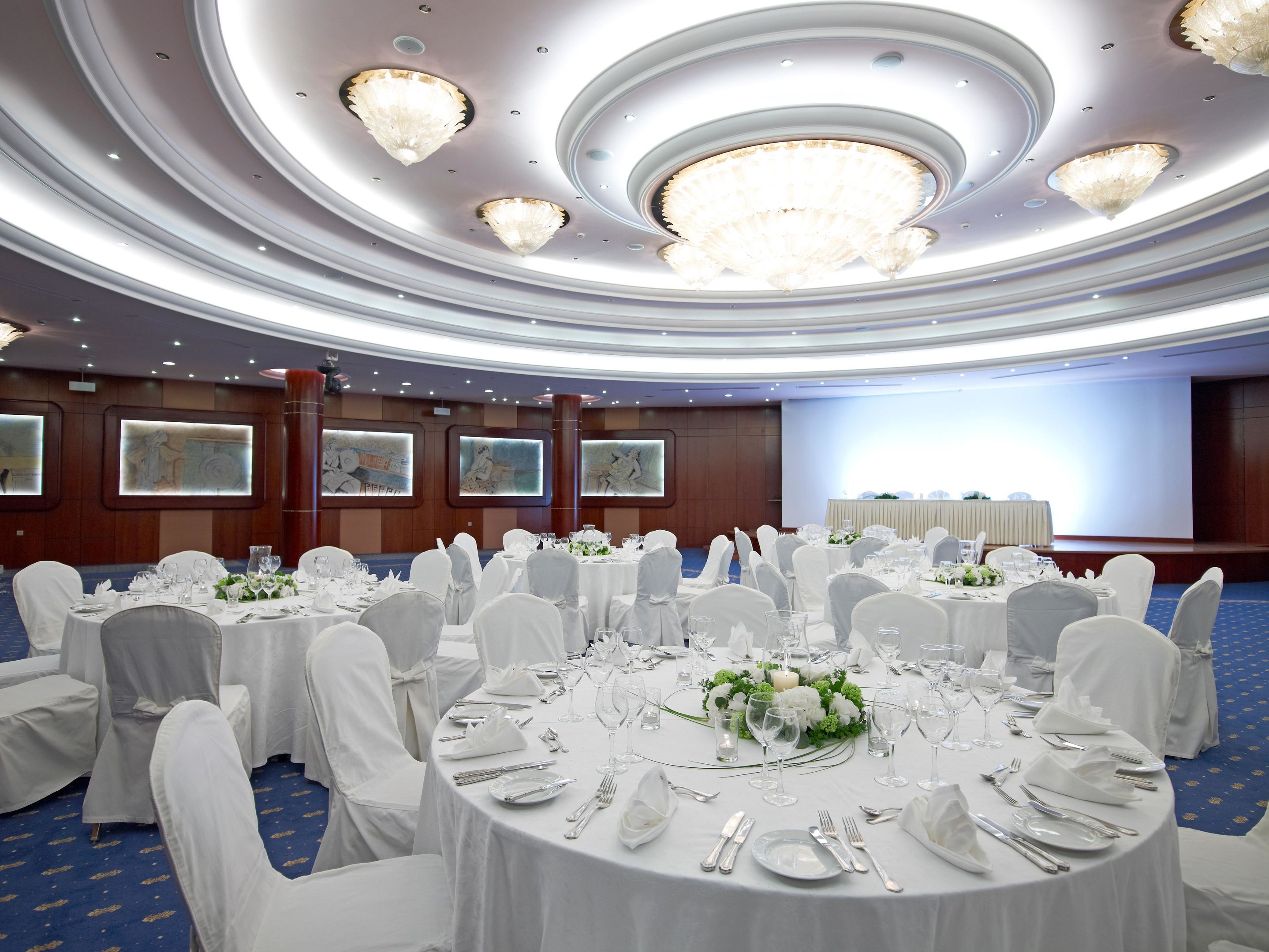 Un salón de banquetes con mesas redondas dispuestas para comer, sillas blancas y candelabros.