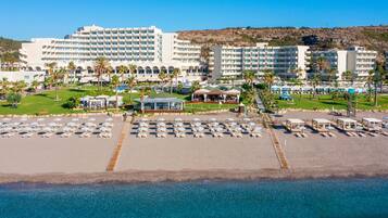 Un resort con varias edificaciones hoteleras, una playa con tumbonas y un área de piscina.