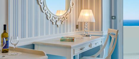 A hotel room with a white desk, a chair, a mirror, a lamp, and a view of the ocean.