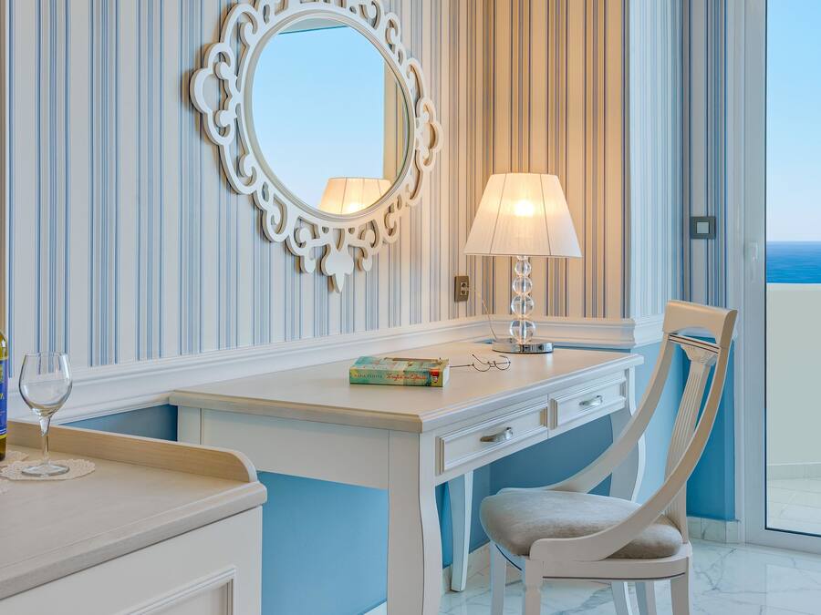 Habitación de hotel con escritorio blanco, una silla, un espejo, una lámpara y vista al océano.