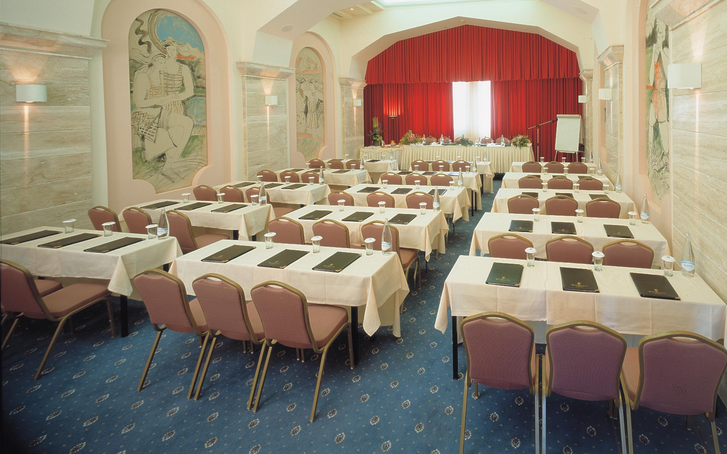 A conference room with rows of tables and chairs, a stage with a red curtain, and decorative wall art.
