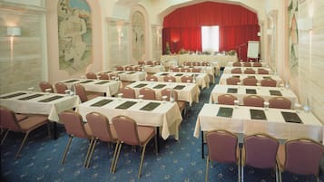 A conference room with rows of tables and chairs, a stage with a red curtain, and decorative wall art.