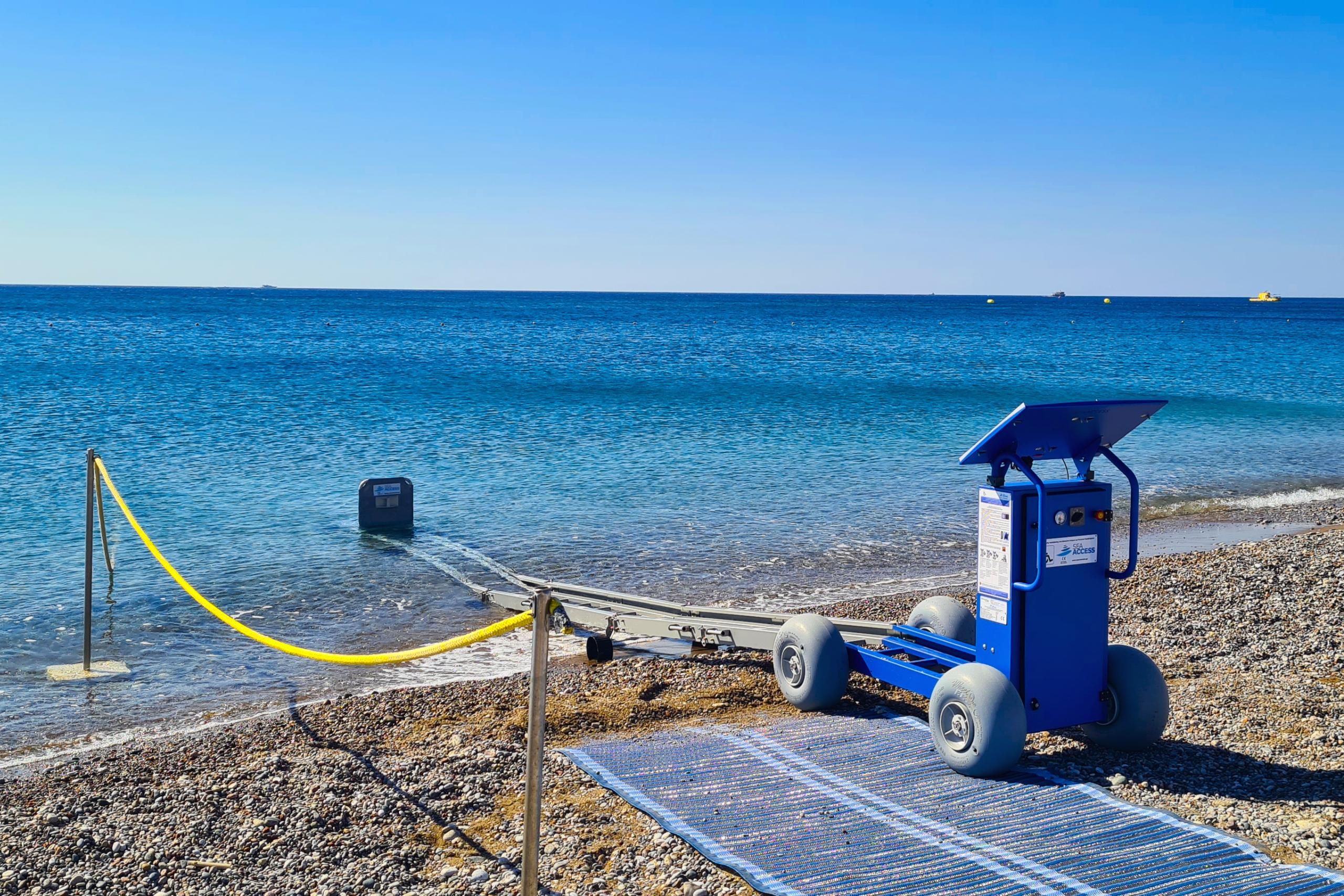 Une plage avec une chaise de maître-nageur bleue, une barrière de corde jaune et un tapis bleu sur le rivage de galets.