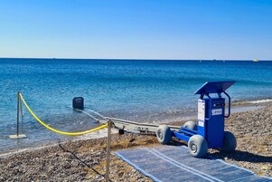 Egy strand, ahol egy kék színű mentőtorony, egy sárga színű, kötelekkel elzárt terület és egy kék színű szőnyeg található a kavicsos parton.