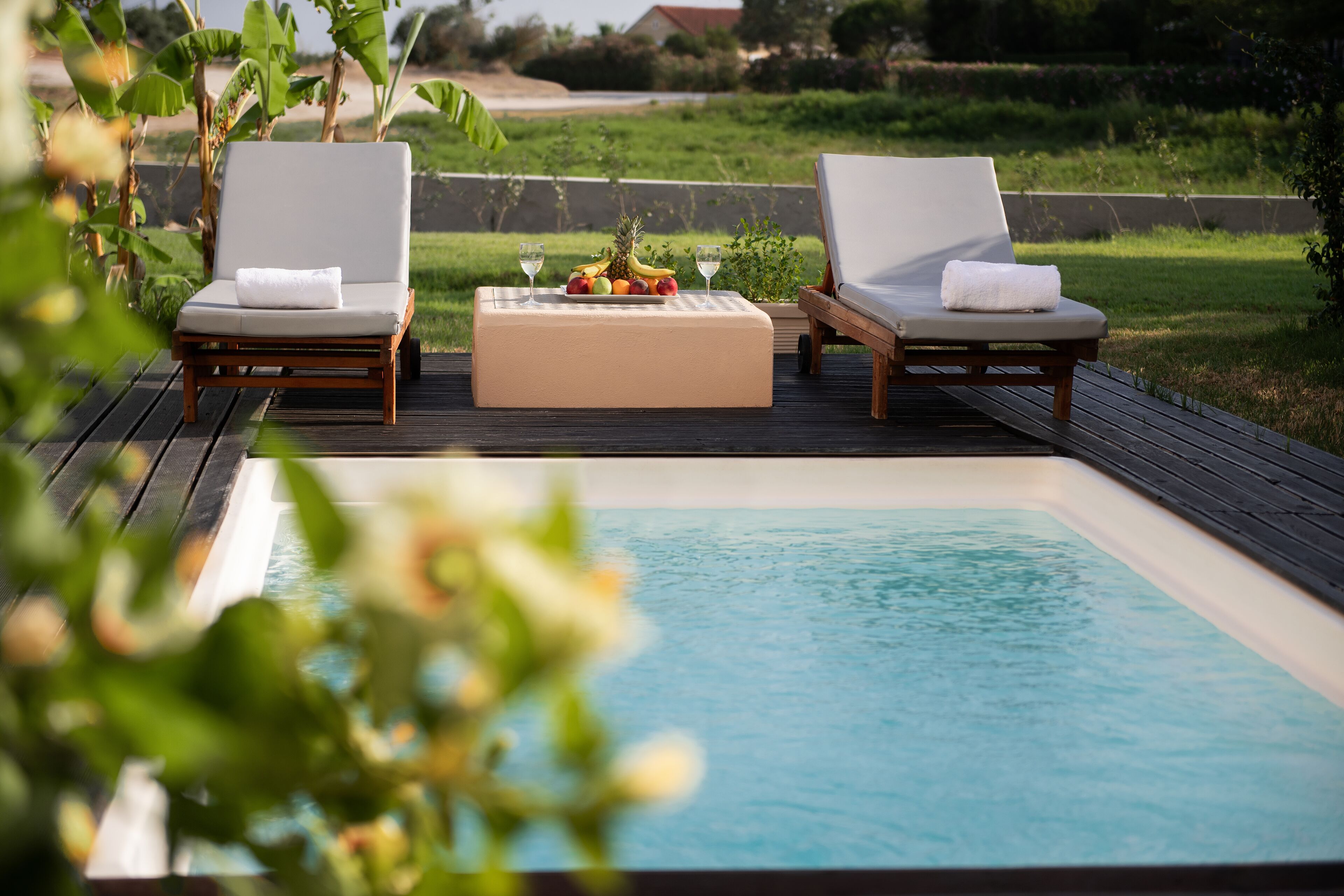 A poolside area with two lounge chairs, a table with fruit and wine glasses, and a view of a lush garden.