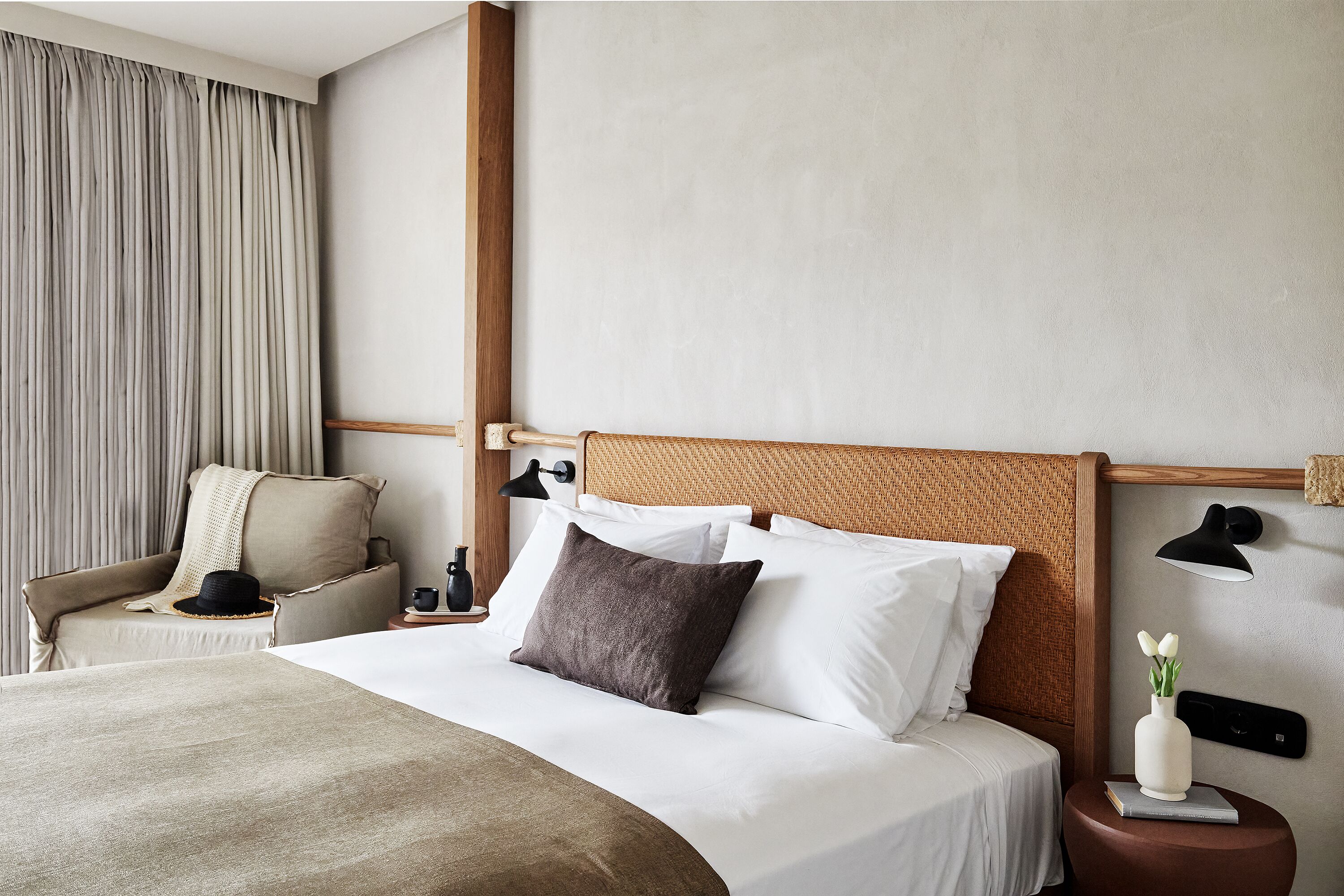 A neatly made bed with white and brown bedding, a wooden headboard, a bedside table with a lamp, and a chair with a hat.