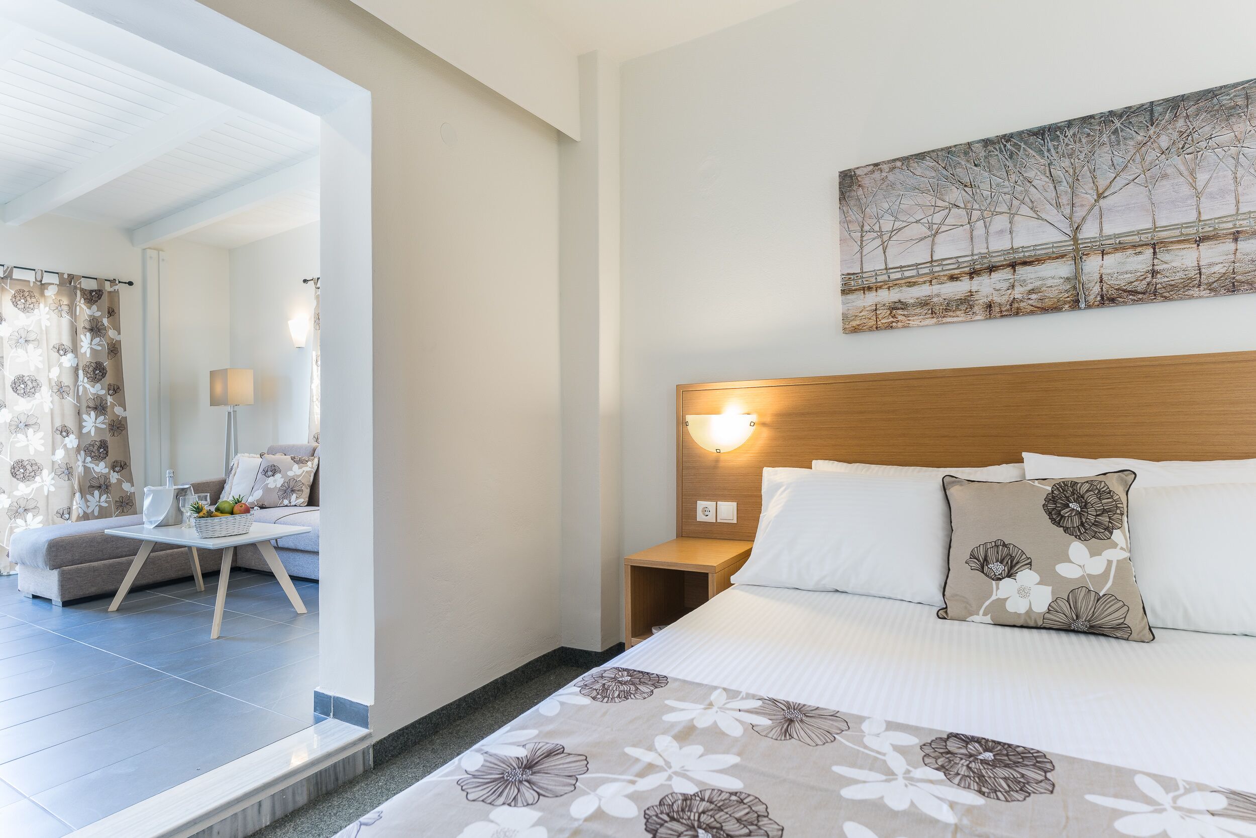 A hotel room with a bed, a nightstand, a lamp, a painting on the wall, and a view of the room through an open door.