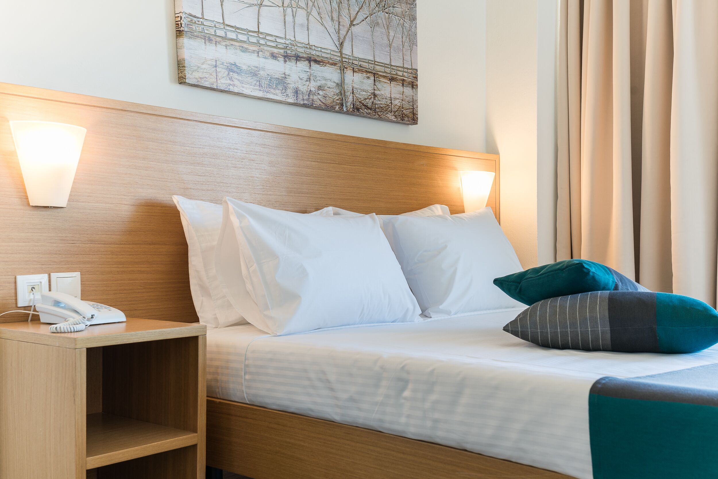 A hotel room with a wooden headboard, a bedside table with a phone, and a painting on the wall.