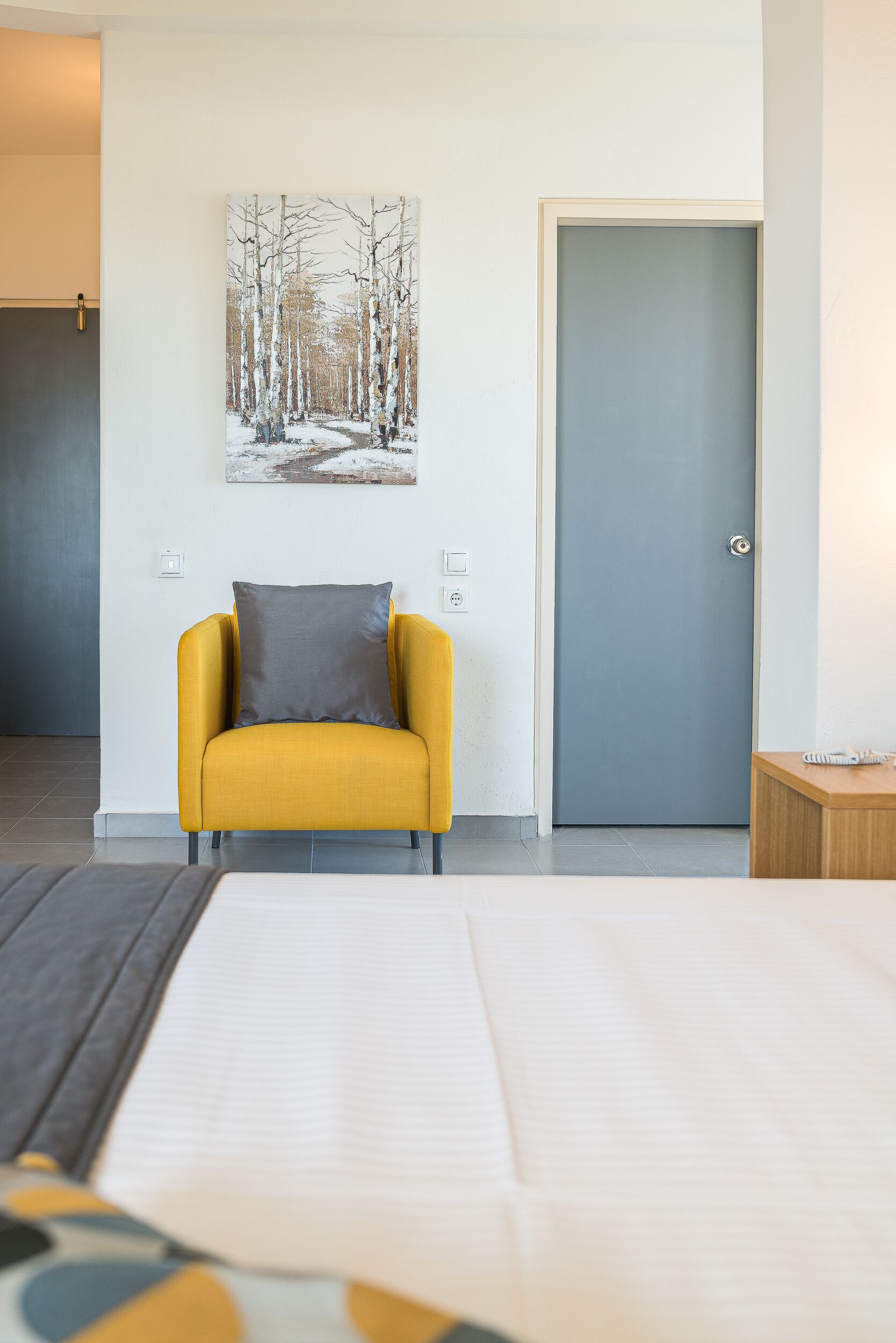 Ein Hotelzimmer mit einem Bett, einem gelben Sessel mit Kissen, einem Bild eines verschneiten Waldes an der Wand und einer geschlossenen Tür.
