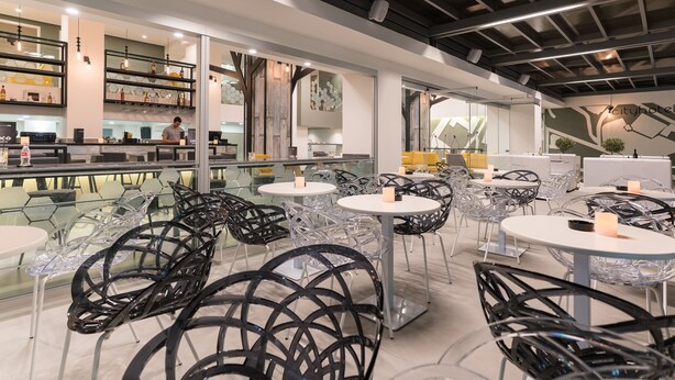 A modern restaurant with glass tables and chairs, a bar area, and a view of the cityscape. - City Center Hotel (Rhodes)