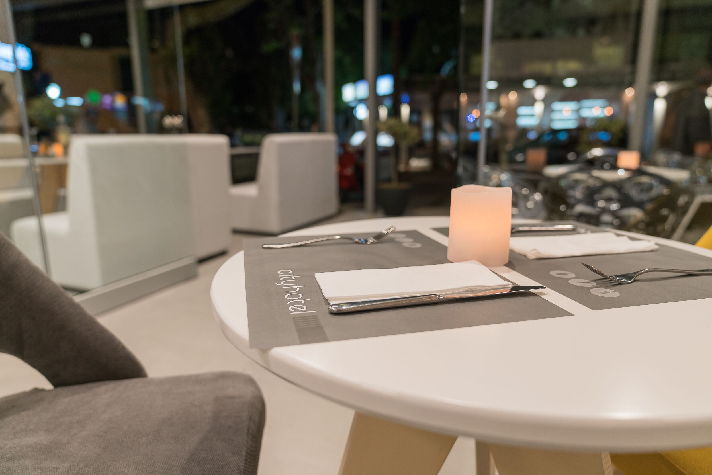 A modern hotel room with a round table, a lit candle, and a view of the city at night.