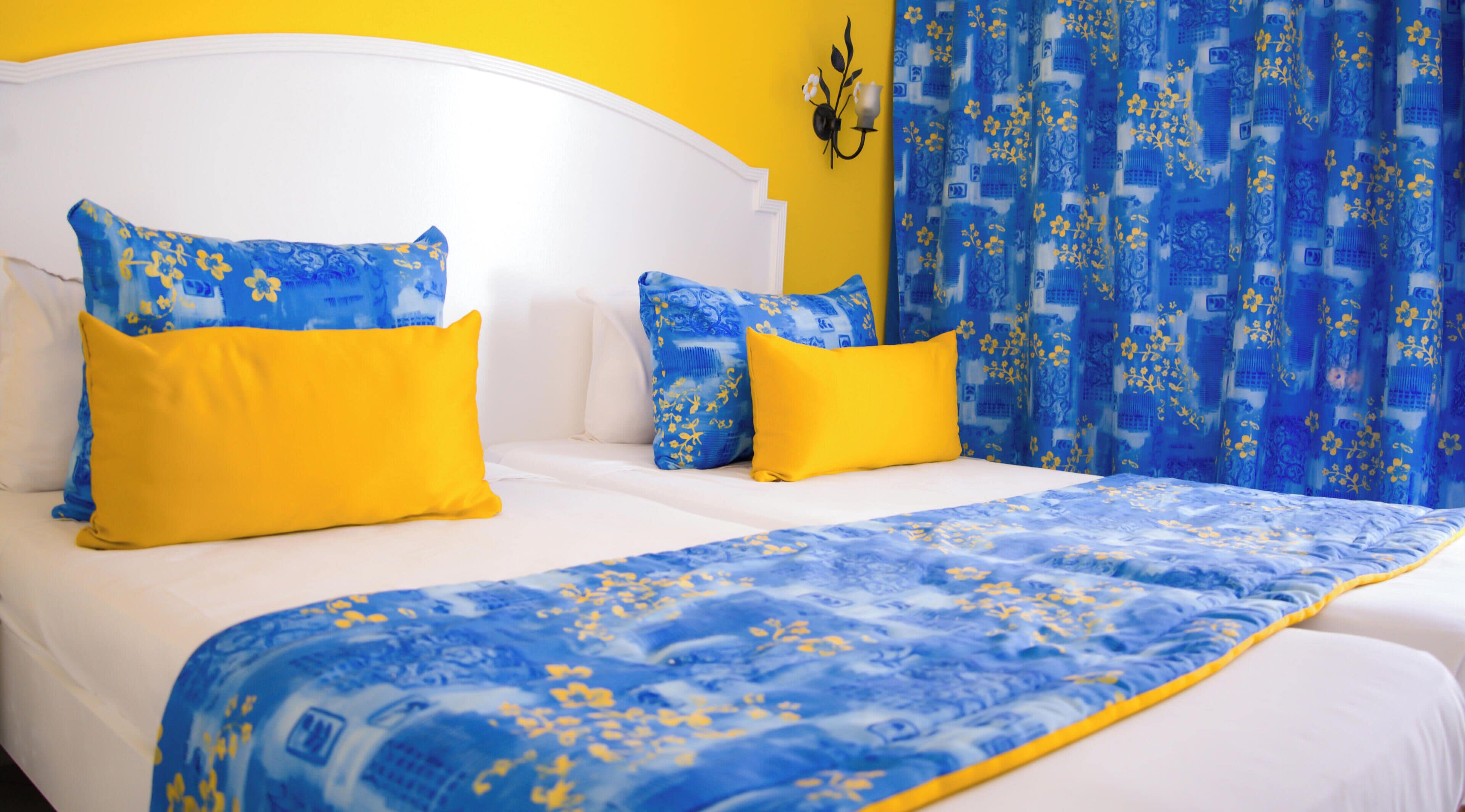 Ein Bett mit weißer Bettwäsche, Überdecken mit blauem Blumenmuster, gelben Kissen und einem blauen Vorhang mit floralem Dekor.