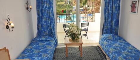 A room with two beds, a small table with a vase, and a view of a pool and palm trees through a glass door.
