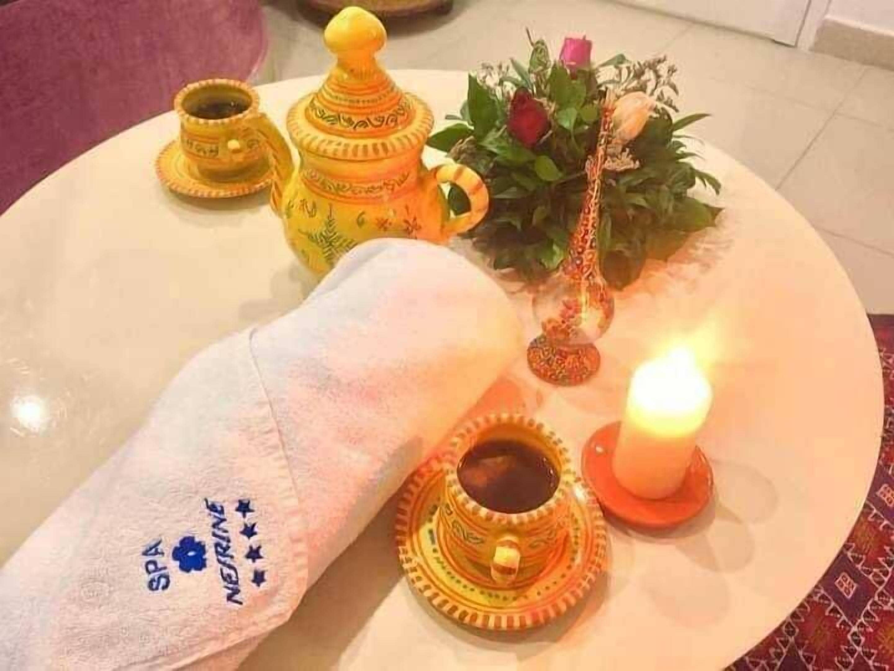 Ein runder Tisch mit weißer Tischdecke, einer brennenden Kerze, einer Zierpflanze und einem Handtuch mit Spa-Logo.
