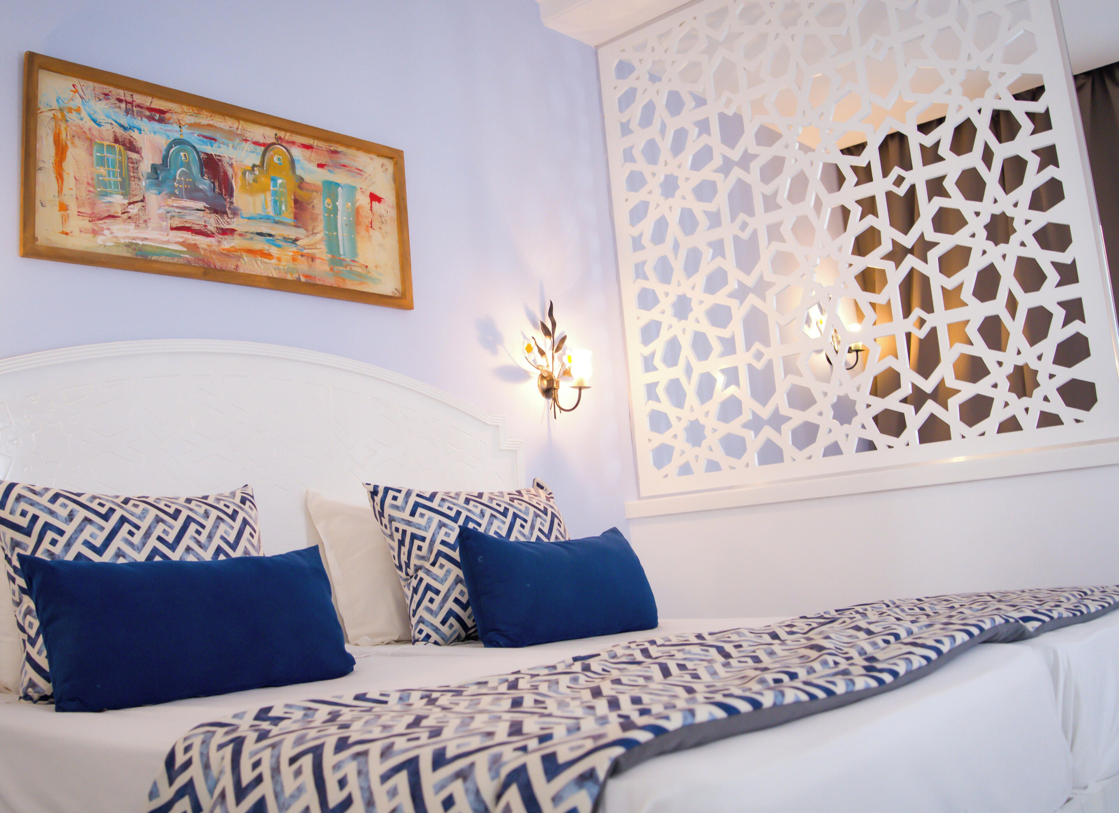 A bedroom with a bed, patterned pillows, a painting, wall sconces, and a decorative screen.