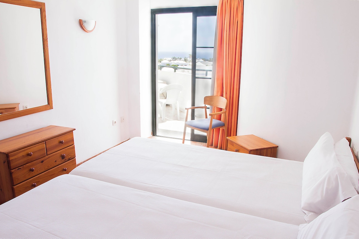 A hotel room with a bed, a wooden dresser, a chair, and a view of the ocean through a sliding glass door.