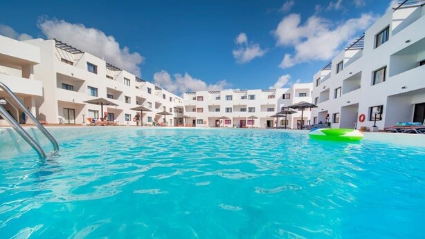 En swimmingpool omgiven av vita byggnader med balkonger, en klarblå himmel och en grön uppblåsbar badleksak.