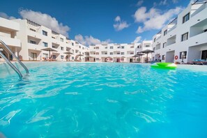Una piscina rodeada de edificios blancos con balcones, cielo azul despejado y un flotador hinchable verde.
