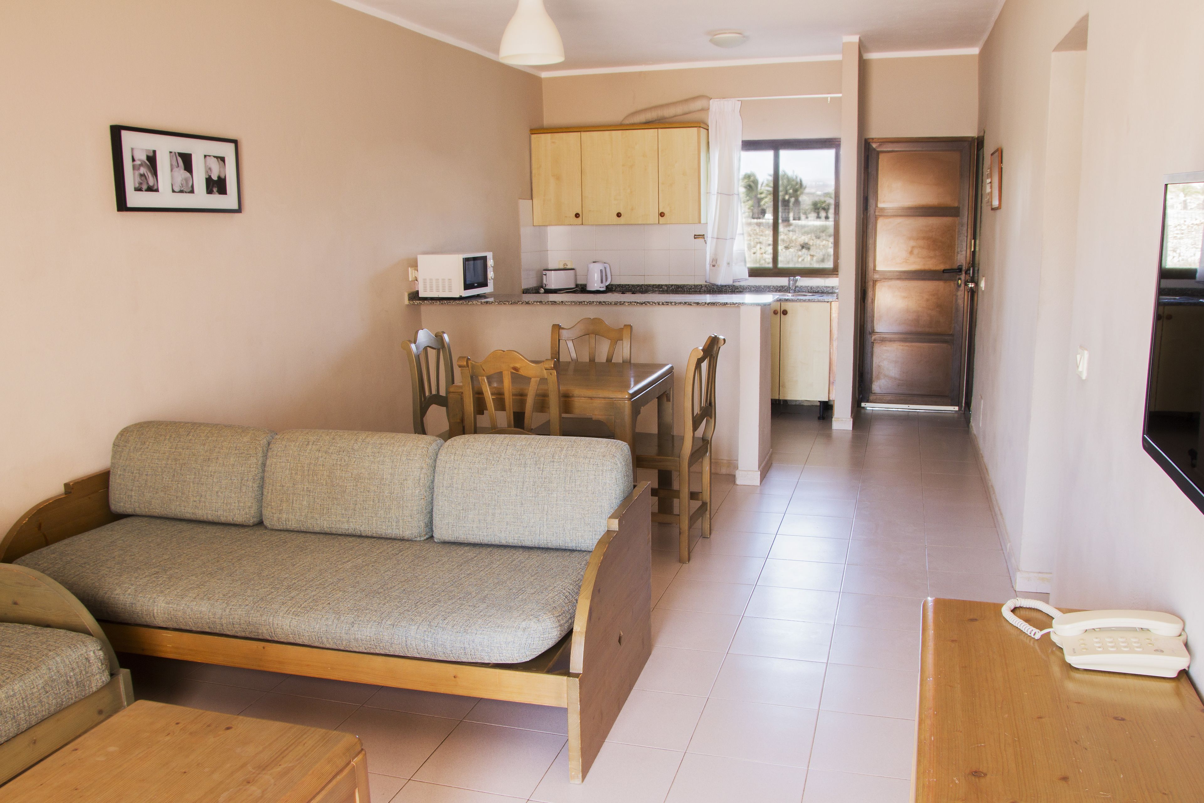 Um espaço de estar compacto com sofá, mesa de jantar, cadeiras, área de cozinha e telefone em uma mesinha lateral.