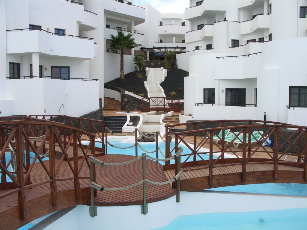 A pool area with wooden walkways and a water slide, surrounded by white buildings with balconies.