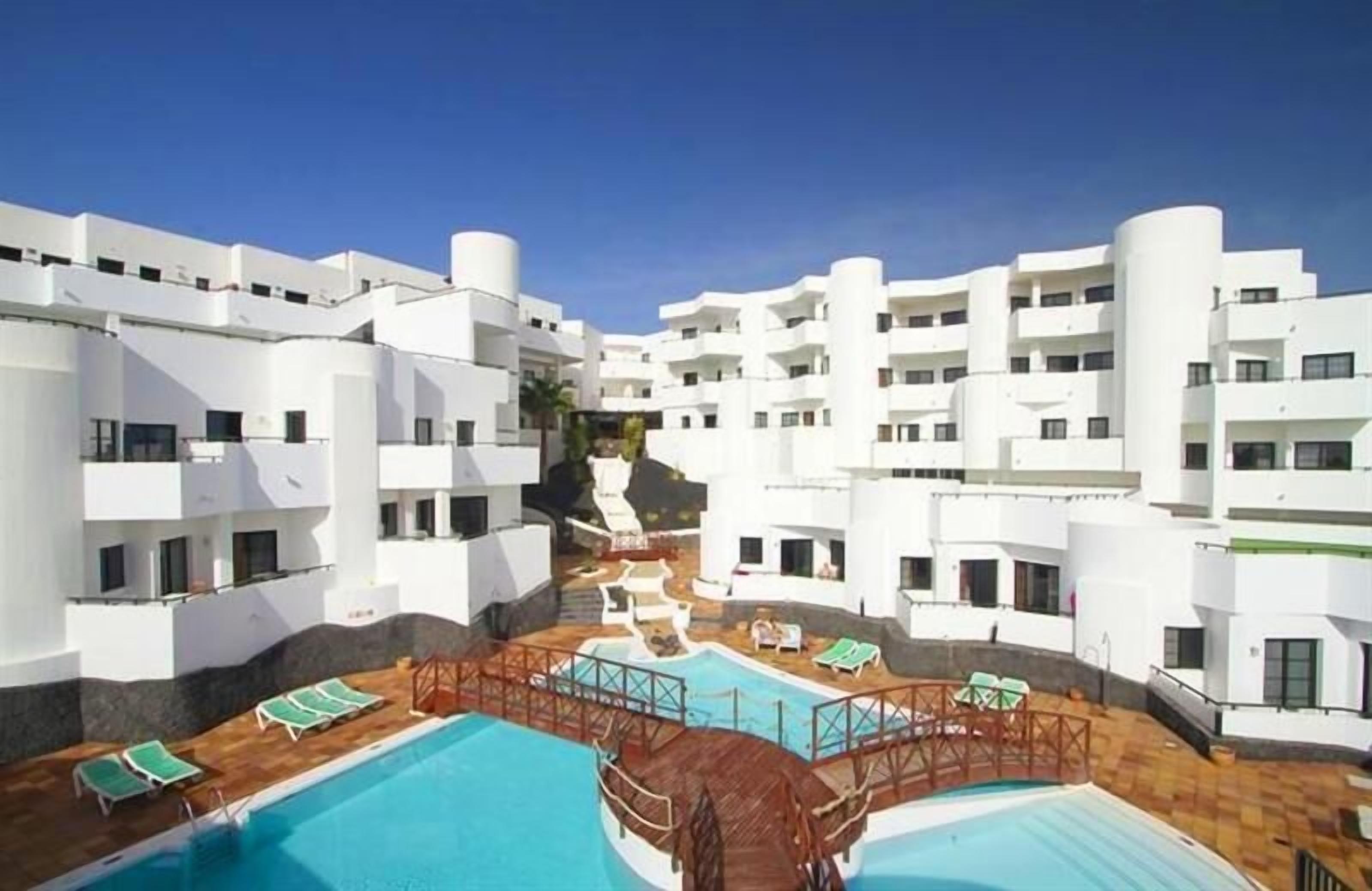 A modern hotel with white exterior walls, a central swimming pool, and multiple balconies.