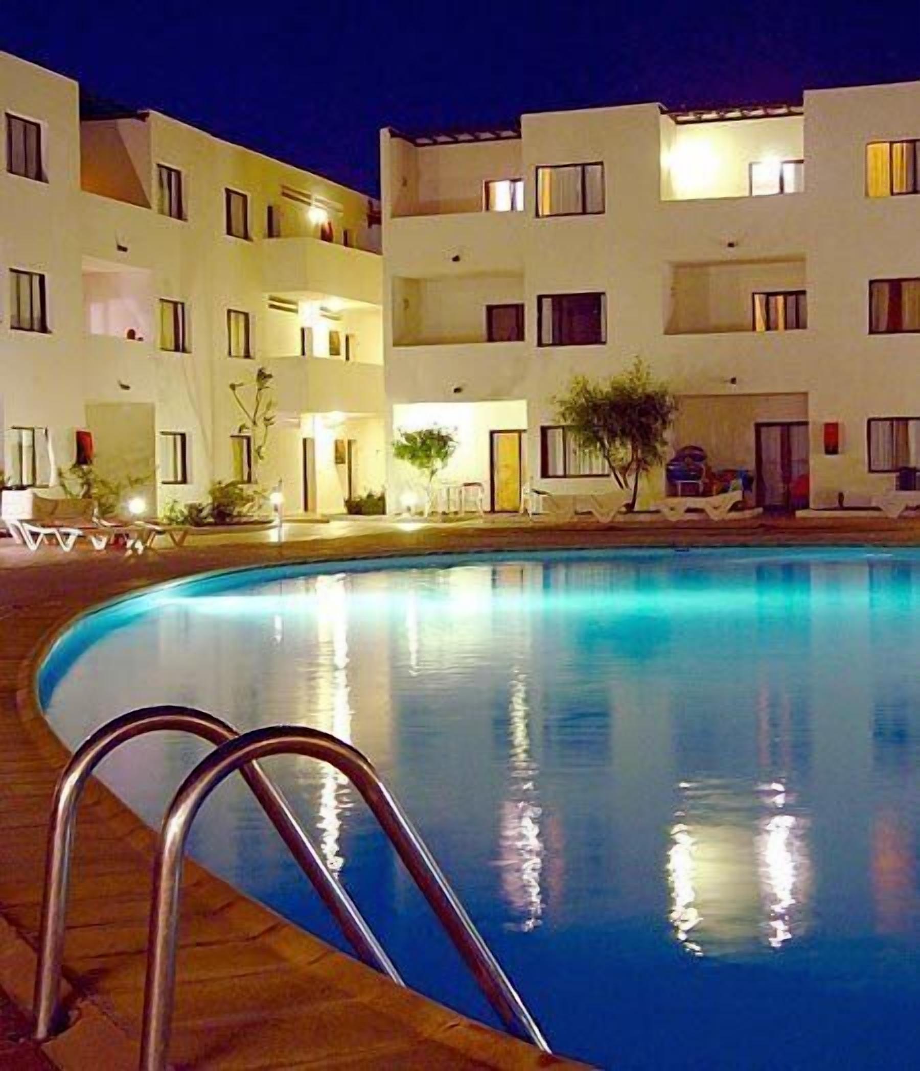 A swimming pool with a wooden deck and metal ladder, surrounded by a multi-story building with balconies and windows.