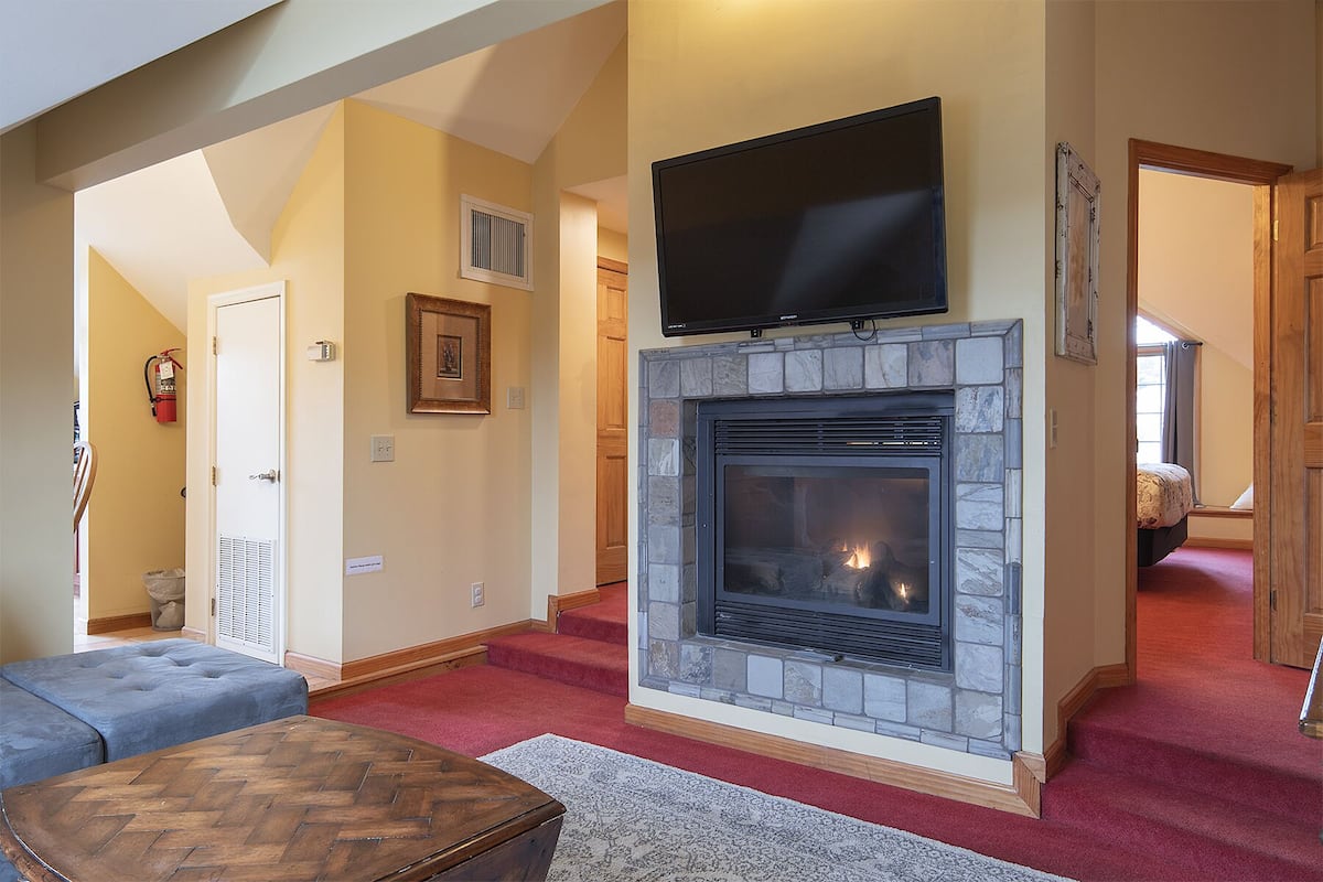A living room with a stone fireplace, a flat-screen TV mounted above it, a blue sofa, and a herringbone-patterned coffee table.