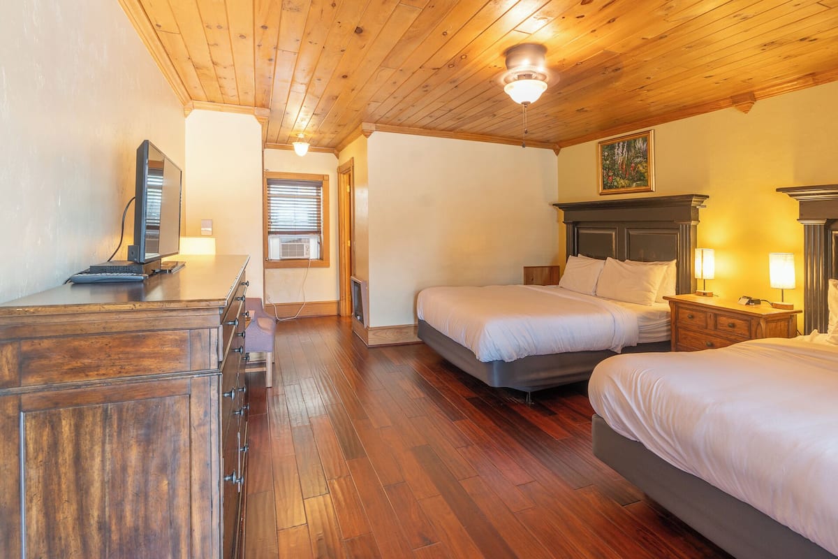 A hotel room with wooden flooring, a bed with a headboard, a desk with a computer, and a window with blinds.