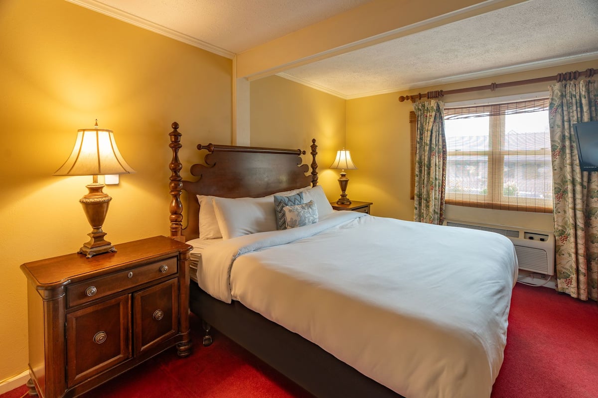 A hotel room with a large bed, two bedside tables with lamps, a window with curtains, and a red carpet.