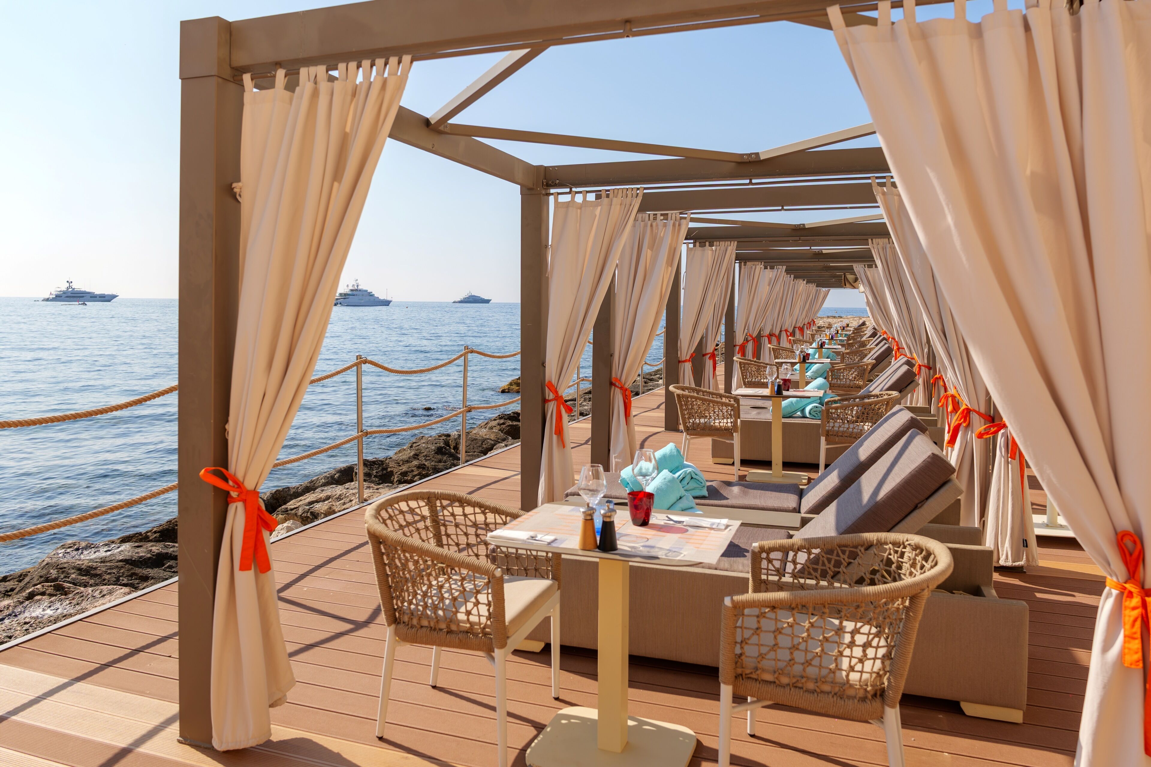 A covered outdoor seating area with wicker furniture, a table set for dining, and a view of the sea with ships in the distance.